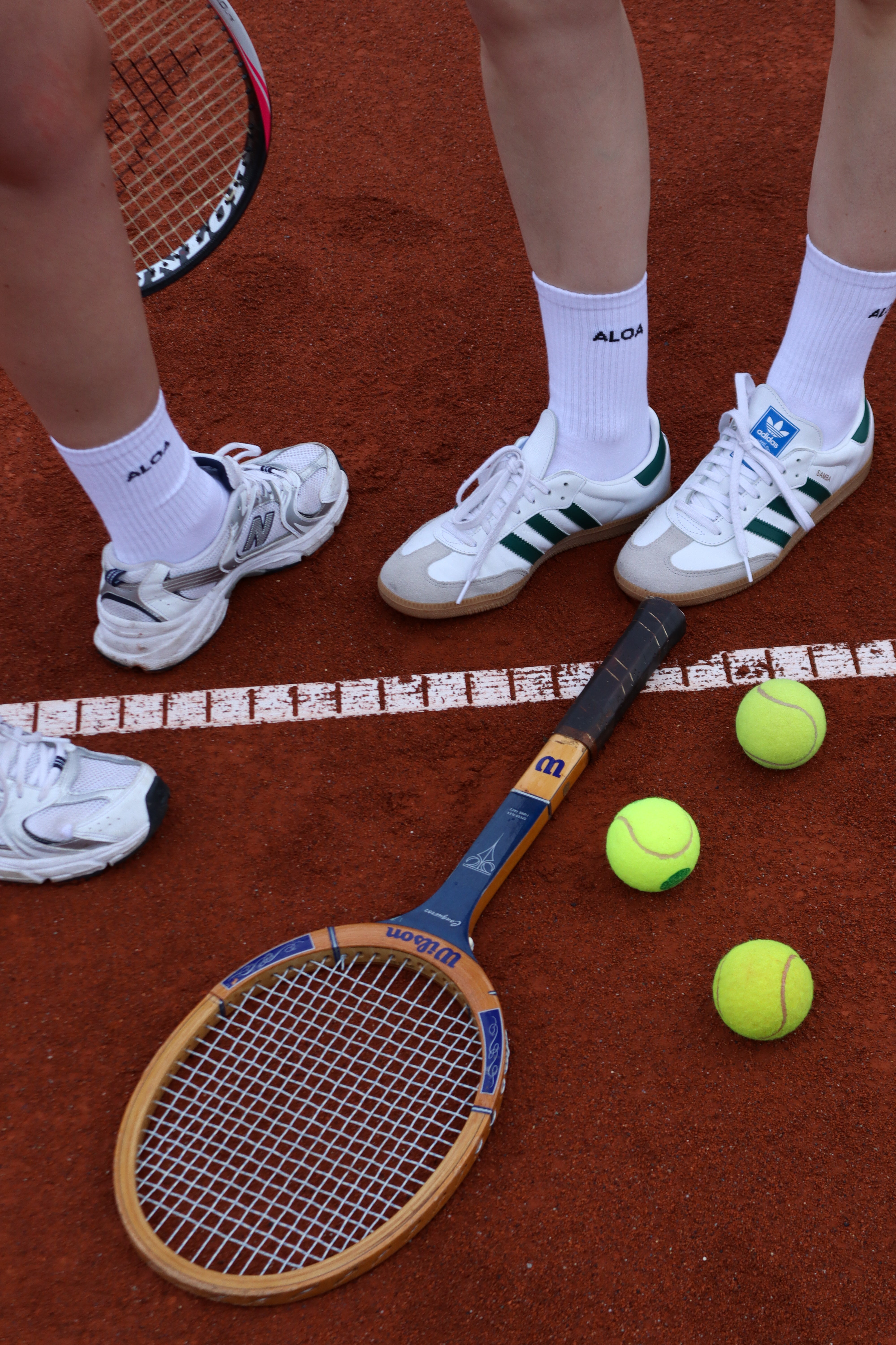 Tennis rackets and balls on a clay court with people wearing white shoes.